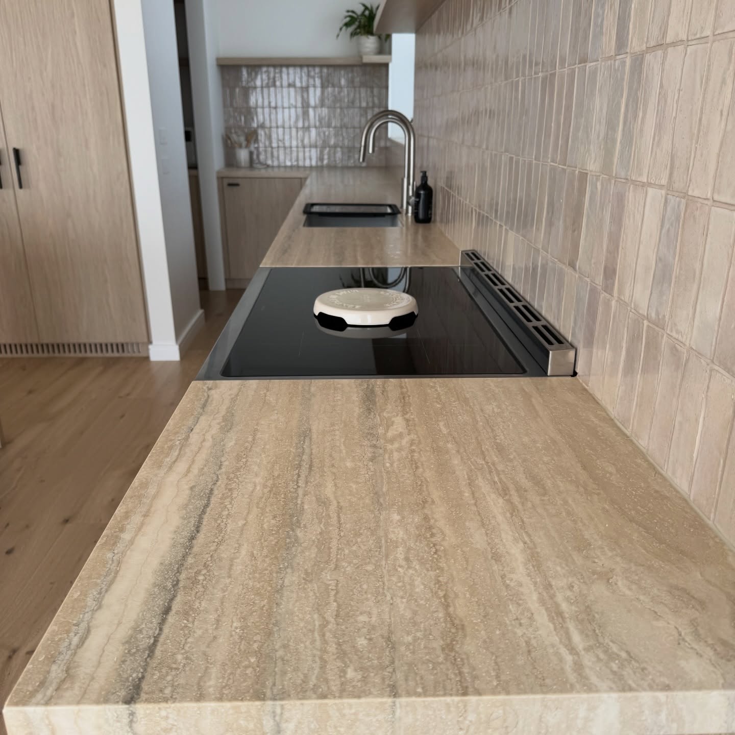 Long travertine benchtop protected with Stone Shield film, running past an induction cooktop to stainless tapware and an undermount sink